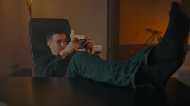 En man spelar i telefonen sitter vid bordet, kastar fötterna på bordet — Stockvideo