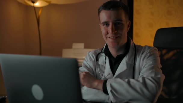 Sonriente médico en bata detrás de la computadora portátil sonriendo a la cámara — Vídeo de stock