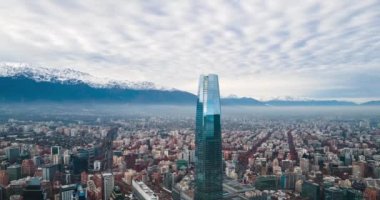 Santiago de Chile şehrinin bulutlu bir gününde, binaları ve kışın karlı sıradağları ile birlikte yüksek hızdaki bir hava manzarası.