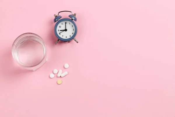 Çalar saat, bir bardak su ve pembe arka planda haplar. Zamanlama.