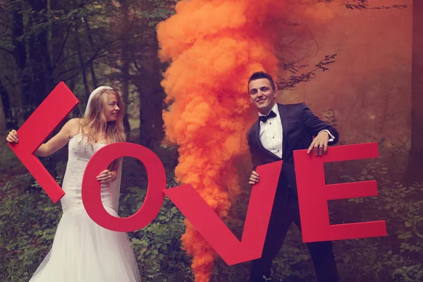 Noiva e noivo segurando grandes letras de amor na floresta, cercado por fumaça vermelha — Fotografia de Stock