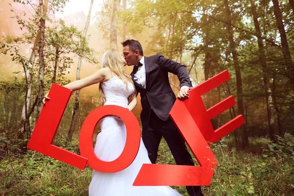 Novia y novio sosteniendo grandes letras de AMOR en el bosque, rodeado de humo rojo — Foto de Stock