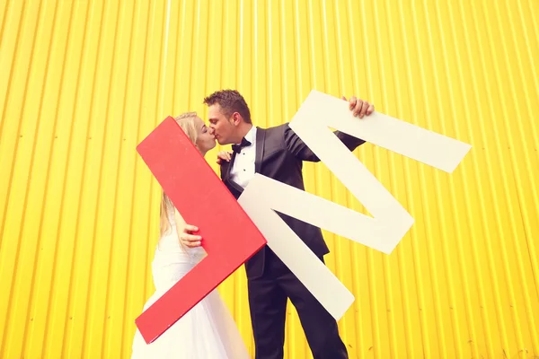 Bride and groom posing against a yellow wall, holding L and M letters — Stock Photo, Image