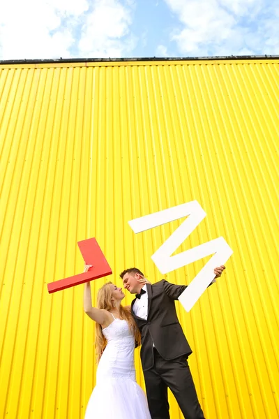 Bruid en bruidegom poseren tegen een gele muur, bedrijf L en M letters — Stockfoto