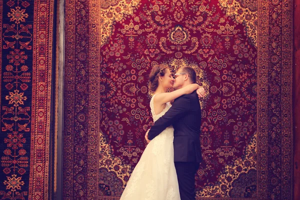 Bride and groom embracing against a hanging carpet — Stock Photo, Image