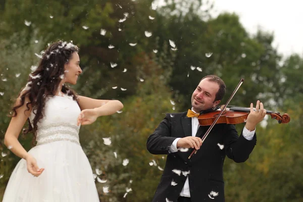 Bela noiva e noivo se divertindo com penas no parque com noivo tocando no violino — Fotografia de Stock