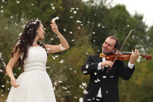Vackra bruden och brudgummen ha kul med fjädrar i park med brudgummen spelar på fiol — Stockfoto