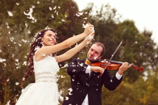 Beautiful bride and groom having fun with feathers in park with groom playing at violin — Stock Photo, Image