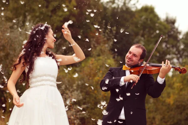 Beautiful bride and groom having fun with feathers in park with groom playing at violin — Stock Photo, Image