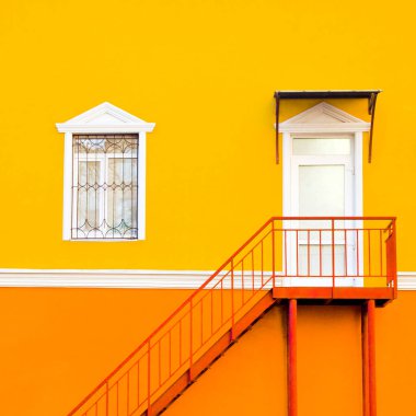 Parlak sarı ve turuncu duvar. İkinci katta penceresi ve girişi olan kırmızı merdivenleri olan bir binanın sulu cephesi. Kare biçimi.