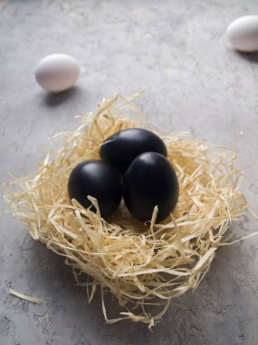 Gri beton bir yüzeydeki yuvada üç siyah yumurta. Arka planda henüz boyanmamış iki beyaz yumurta var. Paskalya için fikirler.