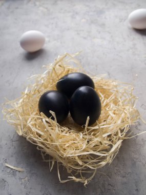 Gri beton bir yüzeydeki yuvada üç siyah yumurta. Arka planda henüz boyanmamış iki beyaz yumurta var. Paskalya için fikirler.