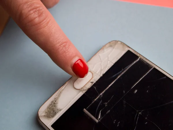 Woman is trying to turn on broken mobile phone. Phone screen is fractured and covered with cracks and sealed with tape. Woman has a sloppy old manicure, which is almost peeled off. Concept of poverty.