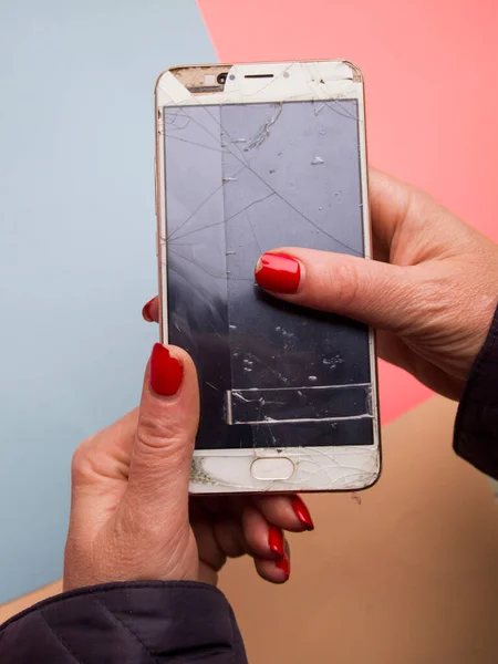 Kırmızı manikürlü bir kadın bozuk bir telefonu tutuyor. Cep telefonunun tamire ihtiyacı var. Ekran bantla kırılmış ve mühürlenmiş. Yıkım ve yoksulluk kavramı.