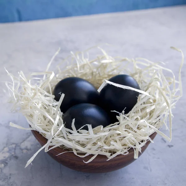 Gri ve mavi beton arka planda samanla dolu killi bir tabakta siyah Paskalya yumurtaları. Geleneksel Paskalya yumurtalarının alışılmadık tasarımı. Kare biçimi.