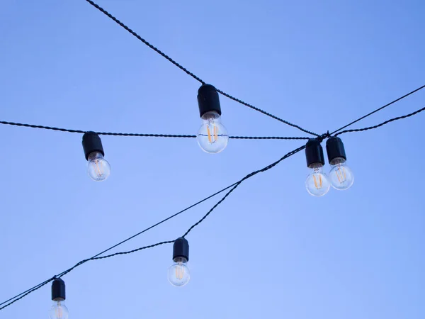 A lot of round light bulbs suspended on a black electric wire. It is a bright sunny day outside and the lights are off.
