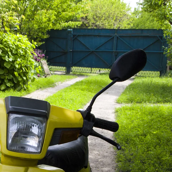 Bahçede güneşli bir gün. Çimen ve ağaçların arka planında siyah plastik dikiz aynası olan sulu sarı bir motosiklet parçası. Seyahat ve macera sembolü. Yaz zamanı. Kare biçimi.