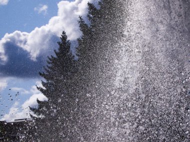 Parktaki çeşmeden tüm yönlere su sıçrıyor. Uzakta koyu renkli kozalaklı siluetler ve beyaz bulutlu parlak mavi bir gökyüzü.