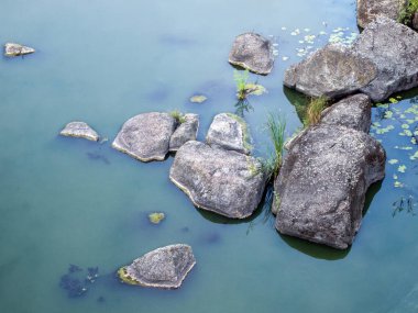 Nehirdeki büyük taşların üst manzarası. Temiz mavi su hafif turbidir ve sütlü bir tonu vardır. Suyun pürüzsüz bembeyaz yüzeyinde çok az yosun ve ördek otu yaprağı vardır..