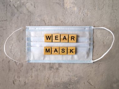Sözcükler, gri bir arka planda beyaz bir cerrahi maskenin üzerine ahşap harflerle kaplanmış Maske takıyor..