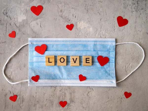 Aşk kelimesi mavi cerrahi maske ve kırmızı kalplerin arka planında ahşap harflerle kaplıydı. Coronavirüs salgını sırasında Sevgililer Günü.