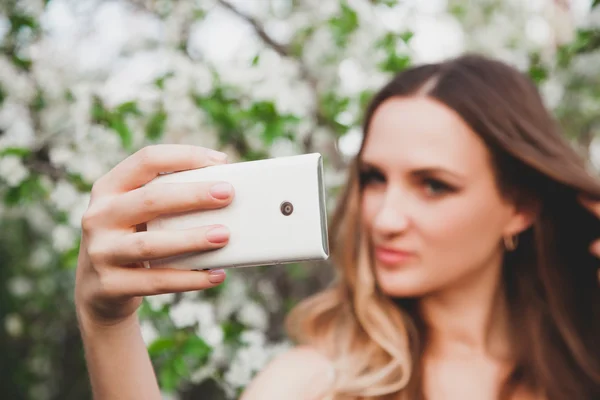 Genç bir kız bir çiçekli ağaç arka plan üzerinde kamera için poz veriyor. O çiçekler yakınındaki selfie alır.