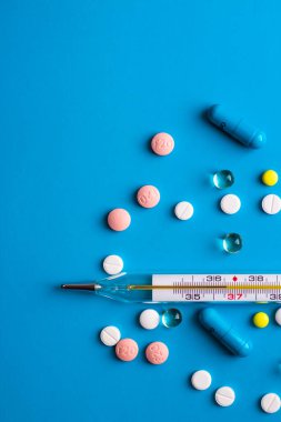 Vücut ısısını ölçmek için civa termometresi, mavi arka planda çok renkli haplar ve kapsüllerle çevrili ve solda yazacak bir yer var. Sağlık ve tedavinin tıbbi teması