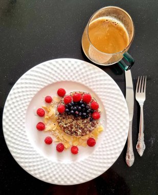 Ahududu, fındık ve fındıkla süslenmiş beyaz bir tabakta kinoa lapası. Yanında çatal bıçak takımı ve masanın üstünde bir fincan kahve olan beyaz bir tabakta sağlıklı ve doğru bir kahvaltı.