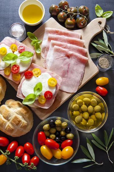 Sándwich con jamón, tomates cherry, aceitunas verdes y negras, albahaca. sobre tabla de madera vieja . —  Fotos de Stock