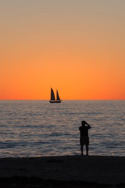 Gün batımında sahilde bir adam ufukta yelkenlinin fotoğrafını çekiyor.. 