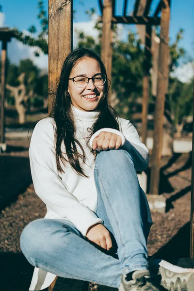 Gözlüklü, beyaz gömlekli ve kot pantolonlu bir kız bankta oturuyor.