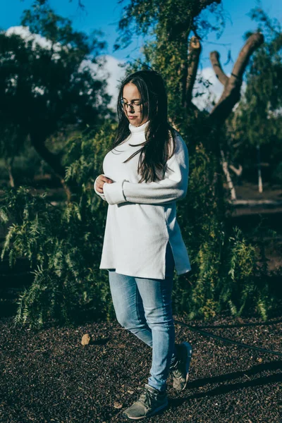 Güneş batarken parkta poz veren gözlüklü, beyaz gömlekli ve kot pantolonlu bir kız.