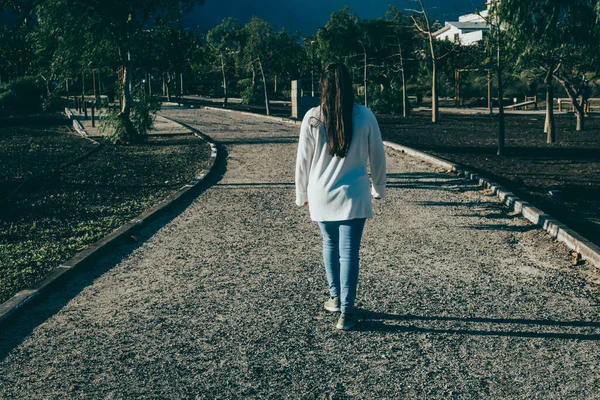 Gözlüklü, beyaz tişörtlü ve kot pantolonlu bir kız parkın ortasında yürüyor.