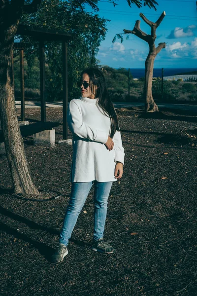 Güneş batarken parkta poz veren gözlüklü, beyaz gömlekli ve kot pantolonlu bir kız.