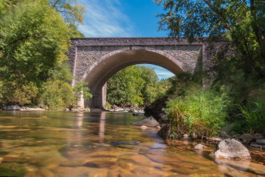 Etrafı ağaçlarla çevrili taş bir köprünün altından geçen nehir.