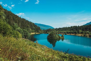 Ormanın ortasında gölün etrafı ağaçlar ve dağlarla çevrili güneş ve bulutlarla dolu bir günde.