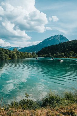 Ormanın ortasında gölün etrafı ağaçlar ve dağlarla çevrili güneş ve bulutlarla dolu bir günde.