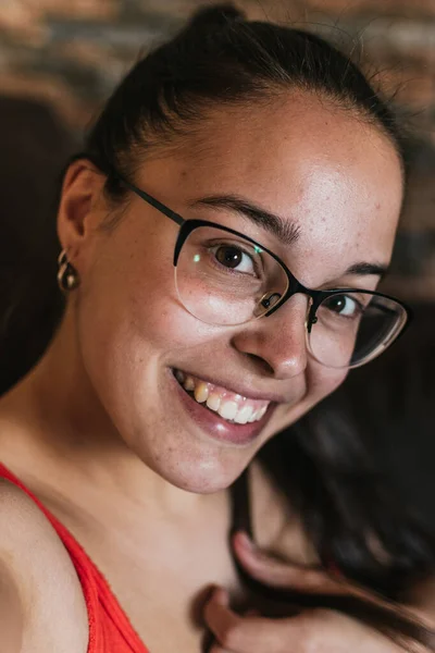 Gözlüklü ve turuncu gömlekli kız kahverengi koltukta video görüşmeleri yapıyor.