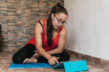 Gözlüklü ve turuncu gömlekli bir kız evde tabletle spor yapıyor.
