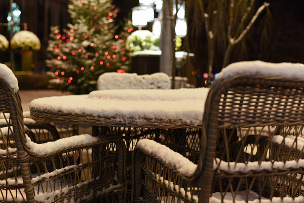 Wicker chairs and table covered with snow in a street cafe on a snowy frosty winter night on Christmas Eve and New Years Eve against the background of festive lighting.