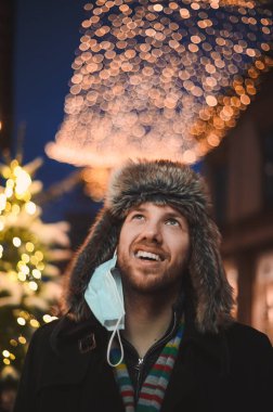 Noel kutlamaları sırasında ve 2021 yılbaşı arifesinde Coronavirus salgını sırasında aydınlanmış şehir ışıklarının arka planına karşı kürk şapkalı çekici bir adam.