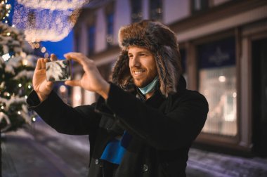 Kürk şapkalı ve yüz maskeli çekici bir adam Noel kutlamaları sırasında ve 2021 yılbaşı arifesinde Coronavirus covid-19 pandemisi sırasında aydınlatılmış şehir ışıkları altında.