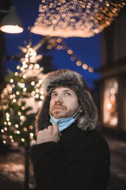Kürk şapkalı ve yüz maskeli çekici bir adam Noel kutlamaları sırasında ve 2021 yılbaşı arifesinde Coronavirus covid-19 pandemisi sırasında aydınlatılmış şehir ışıkları altında.