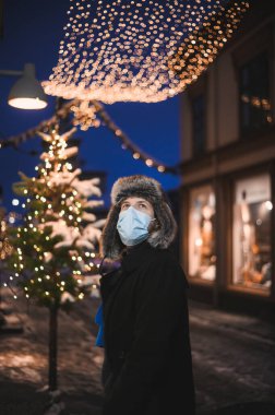 Kürk şapkalı ve yüz maskeli çekici bir adam Noel kutlamaları sırasında ve 2021 yılbaşı arifesinde Coronavirus covid-19 pandemisi sırasında aydınlatılmış şehir ışıkları altında.