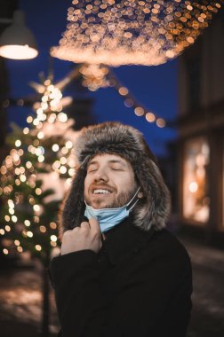 Kürk şapkalı ve yüz maskeli çekici bir adam Noel kutlamaları sırasında ve 2021 yılbaşı arifesinde Coronavirus covid-19 pandemisi sırasında aydınlatılmış şehir ışıkları altında.