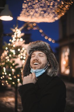 Kürk şapkalı ve yüz maskeli çekici bir adam Noel kutlamaları sırasında ve 2021 yılbaşı arifesinde Coronavirus covid-19 pandemisi sırasında aydınlatılmış şehir ışıkları altında.