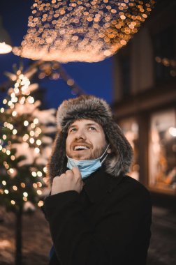 Kürk şapkalı ve yüz maskeli çekici bir adam Noel kutlamaları sırasında ve 2021 yılbaşı arifesinde Coronavirus covid-19 pandemisi sırasında aydınlatılmış şehir ışıkları altında.