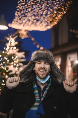 Kürk şapkalı ve yüz maskeli çekici bir adam Noel kutlamaları sırasında ve 2021 yılbaşı arifesinde Coronavirus covid-19 pandemisi sırasında aydınlatılmış şehir ışıkları altında.