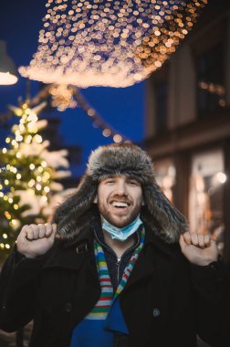 Kürk şapkalı ve yüz maskeli çekici bir adam Noel kutlamaları sırasında ve 2021 yılbaşı arifesinde Coronavirus covid-19 pandemisi sırasında aydınlatılmış şehir ışıkları altında.