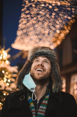 Kürk şapkalı ve yüz maskeli çekici bir adam Noel kutlamaları sırasında ve 2021 yılbaşı arifesinde Coronavirus covid-19 pandemisi sırasında aydınlatılmış şehir ışıkları altında.